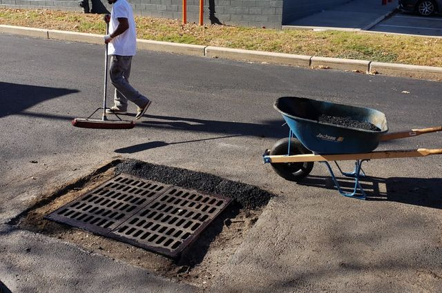 Worker with wheelbarrow and shovel repairing a street near an open storm drain.
