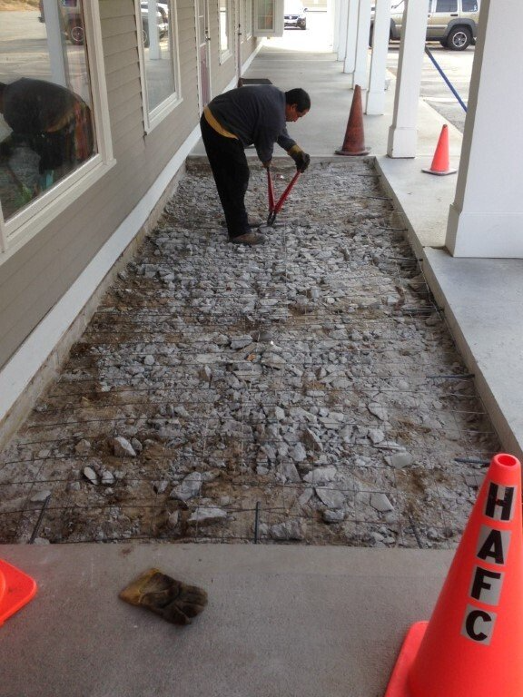 Worker tamping gravel in a walkway beside a building, with orange cones marking the area