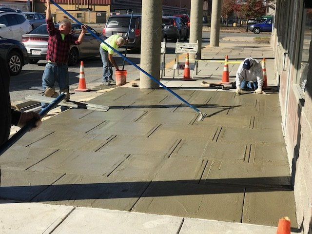Workers smoothing fresh concrete on a sidewalk beside traffic cones and a building