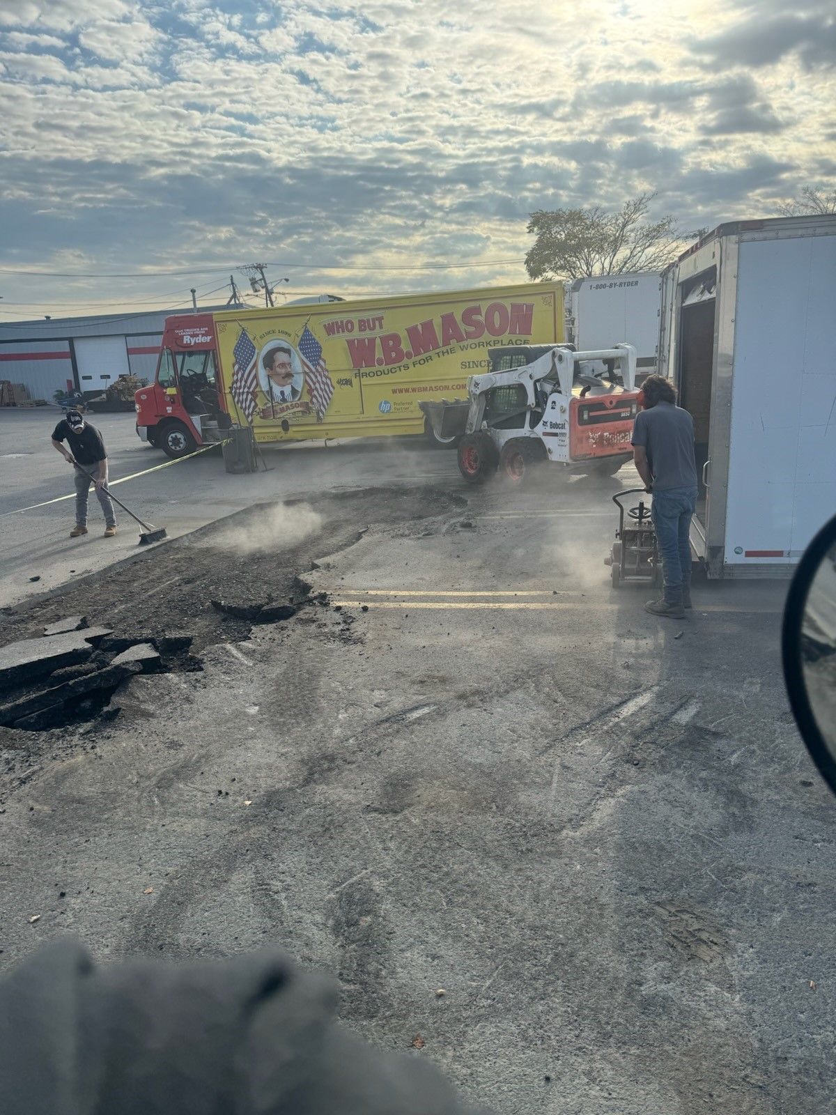 Damaged road with debris, workers, and a yellow truck stopped near a roadside building at sunset.