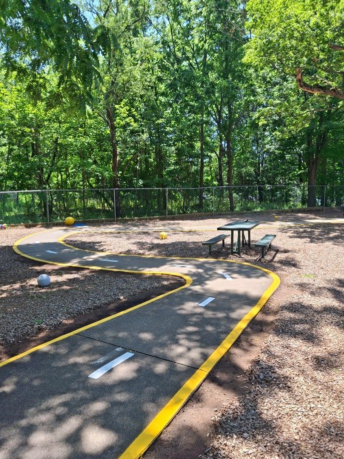Mini golf course with winding gray path, yellow edges, and benches among trees in a wooded park