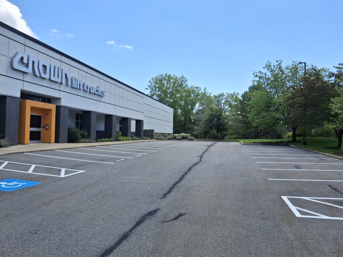 Empty parking lot beside a low white building under a blue sky, with trees in the background.