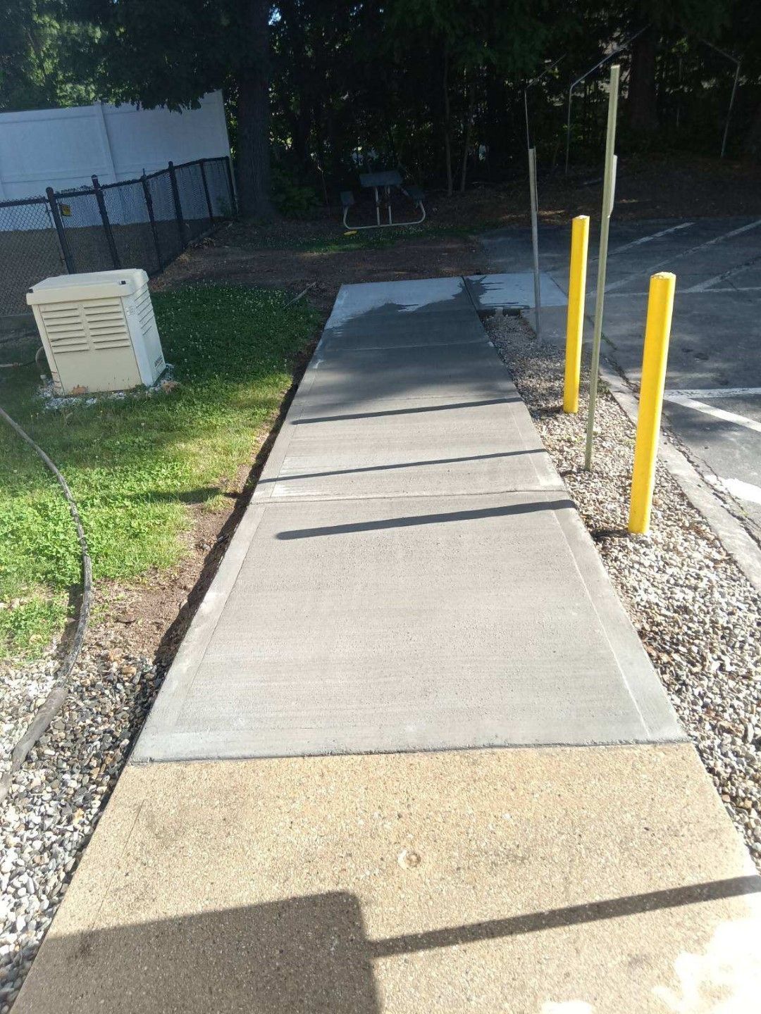 Concrete sidewalk with yellow bollards beside a grassy area and building wall, lit by sunlight and shadow.