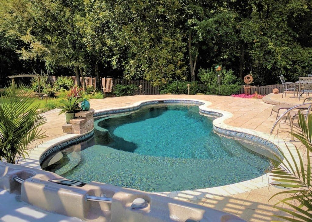 Backyard swimming pool with turquoise water, beige deck, and lush green trees surrounding it