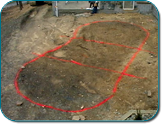 A dirt lot marked with red spray paint outlining several connected shapes