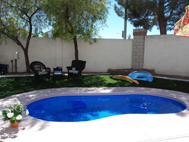 Backyard patio with a small blue pool, chairs, and potted flowers under trees and a white wall