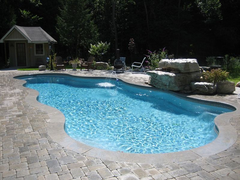 Backyard swimming pool with blue water, stone patio, and a small pool house surrounded by trees