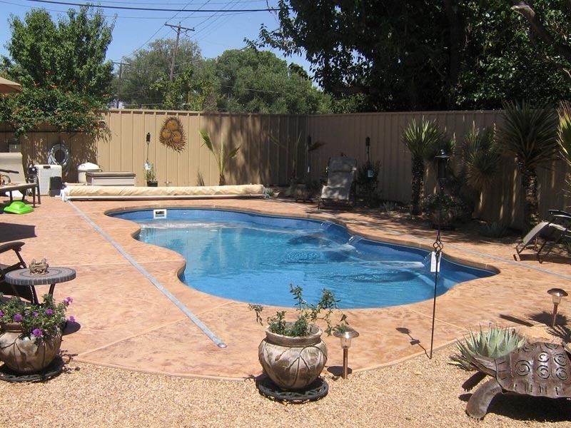 Backyard swimming pool with tan patio, potted plants, wooden fence, and trees on a sunny day