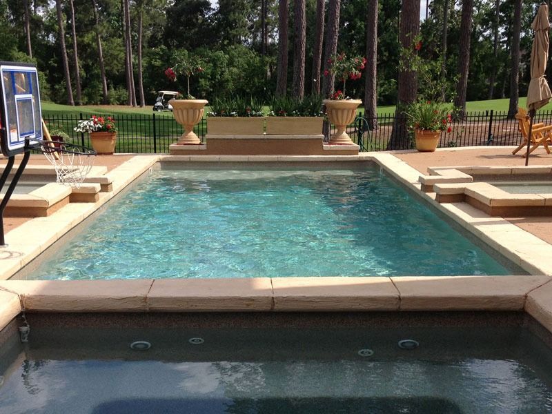 Rectangular turquoise swimming pool bordered by stone, with potted plants and patio chairs in a sunny garden.