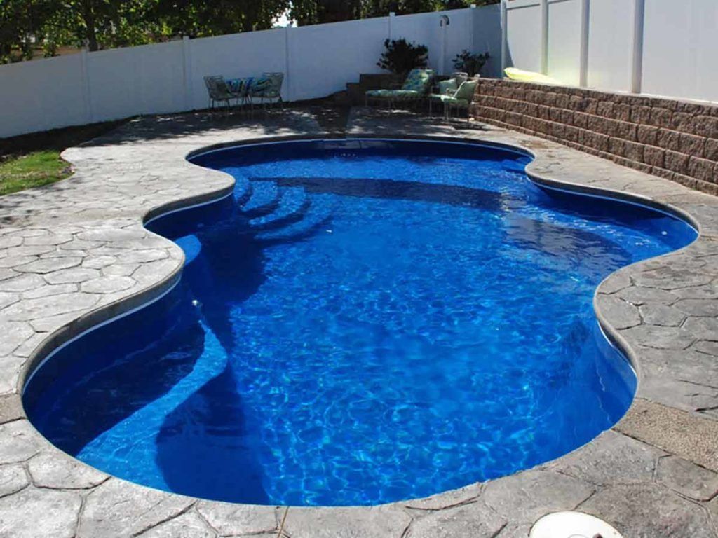 Curved blue swimming pool beside a stone patio and white fence in a sunny backyard