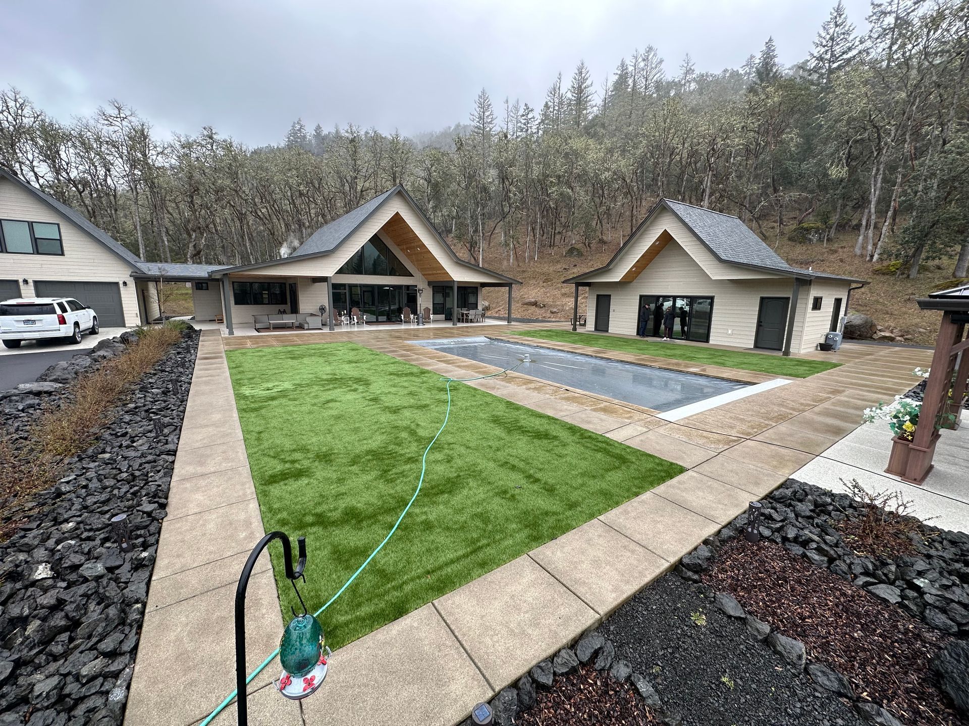Backyard with green lawn, paved path, and two modern small houses on a wooded hillside