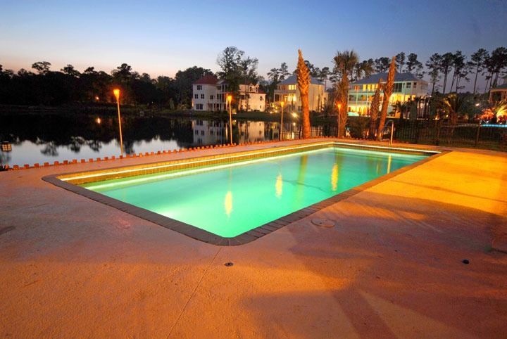 Outdoor swimming pool glowing turquoise at dusk, with warm lights and a lakeside backdrop