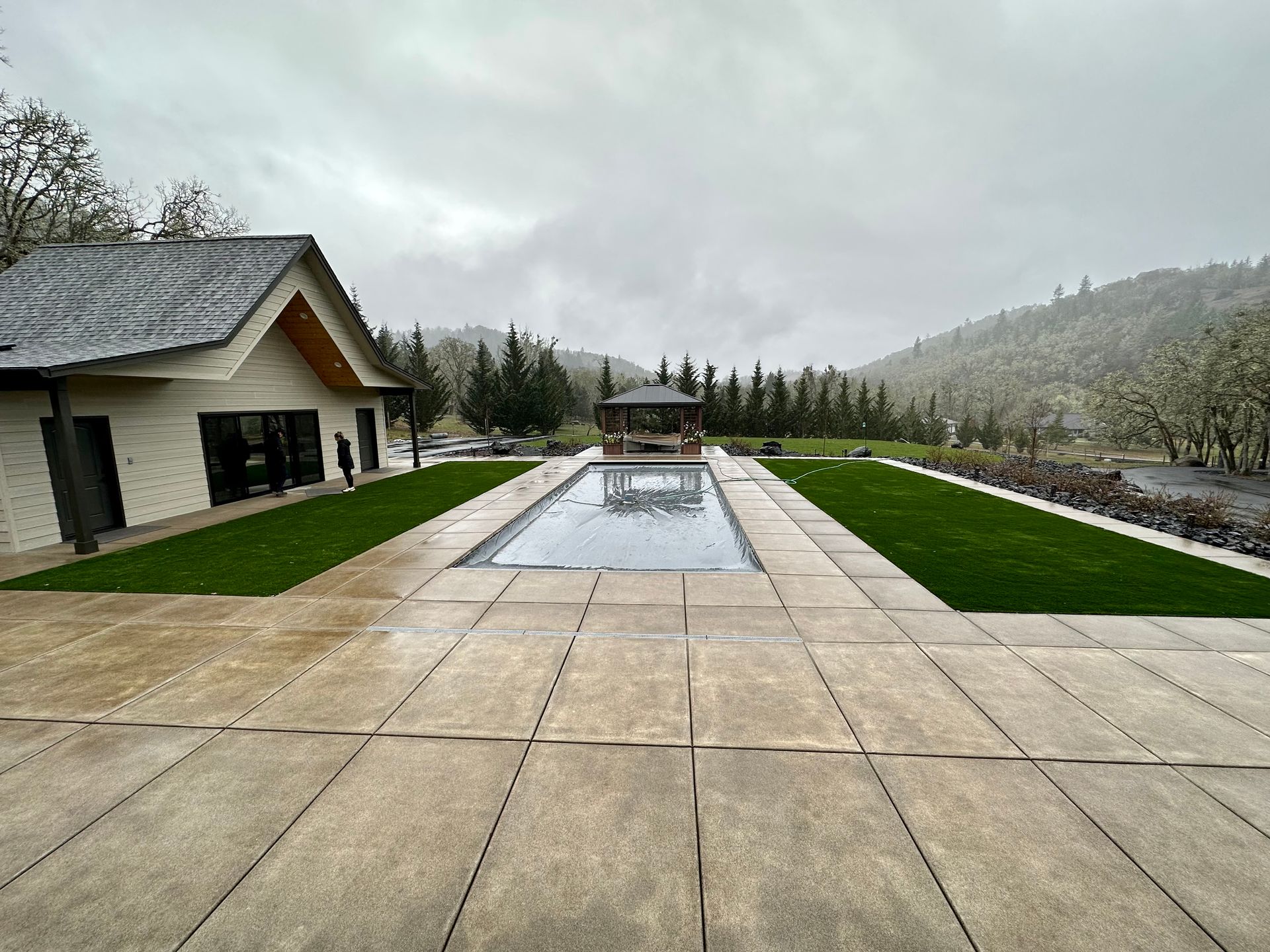 Modern patio with long reflecting pool, green lawns, and misty mountains under a cloudy sky