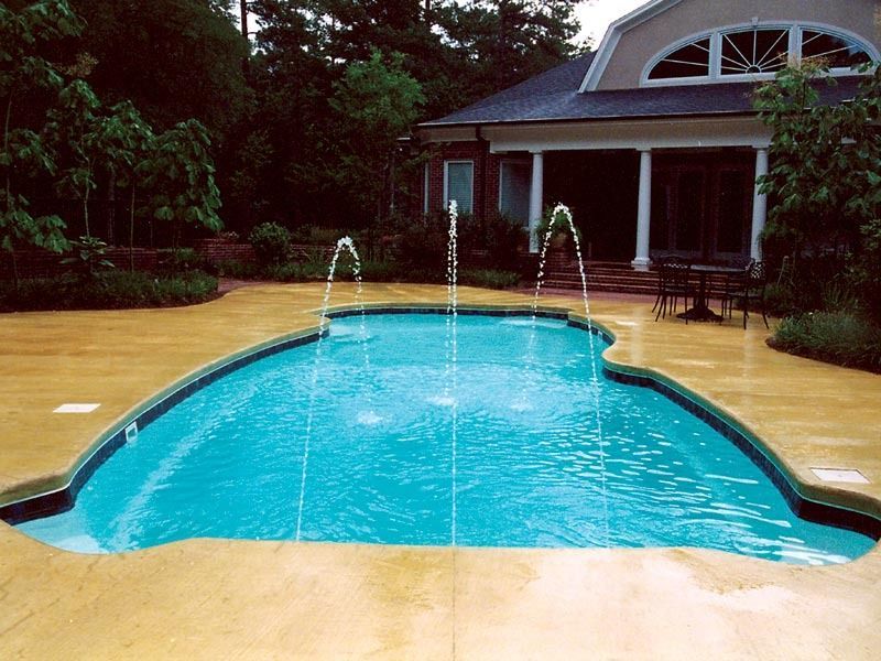 Curved blue backyard pool with three fountain jets in front of a house and trees