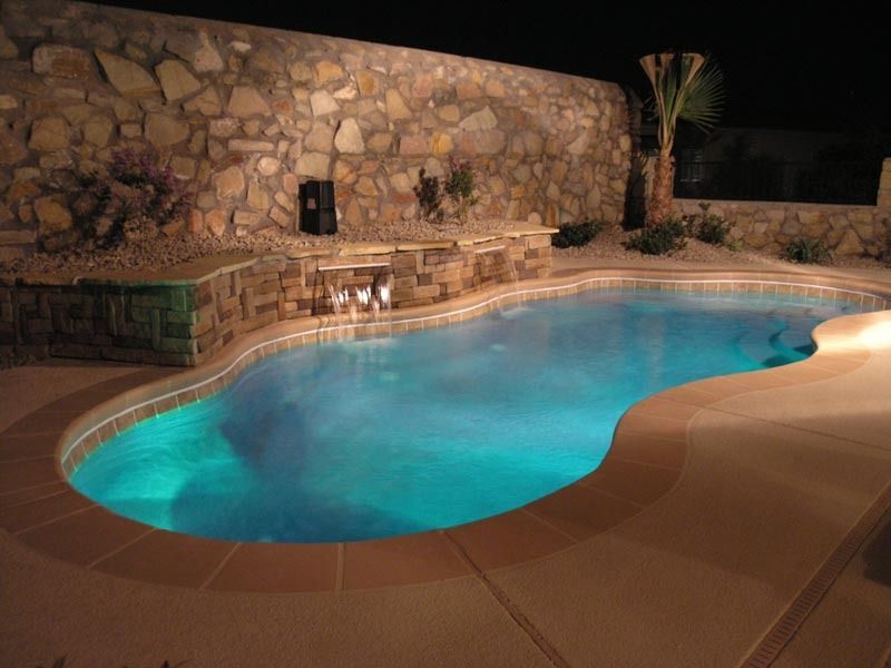 Lit backyard pool glowing blue at night beside a stone wall and patio.