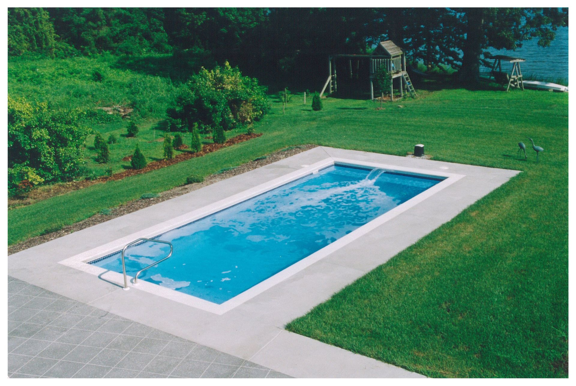 Rectangular outdoor swimming pool with blue water, white deck, and green lawn in a backyard garden