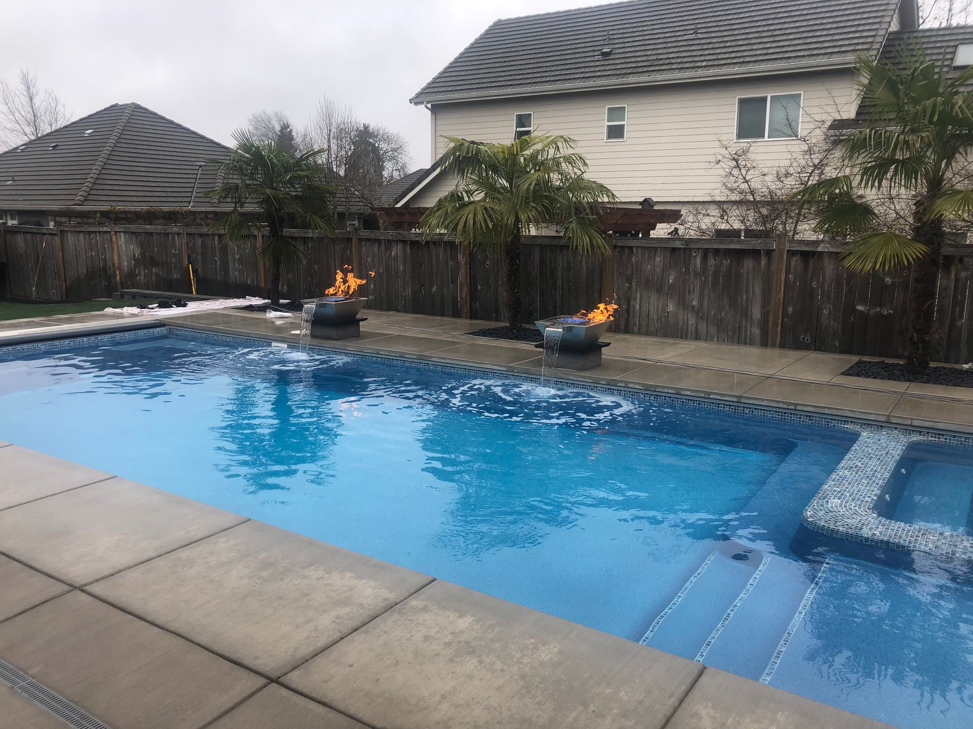 Backyard swimming pool with blue water, stone deck, and two fire bowls by a wooden fence