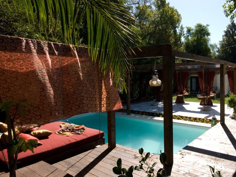 Tropical patio with turquoise pool, red daybed, palm trees, and hanging lanterns beside a modern villa