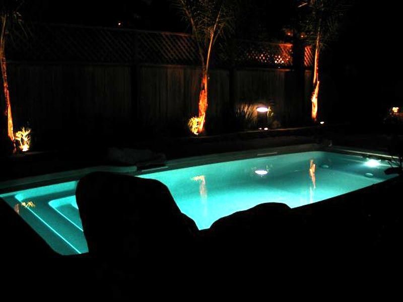 Dimly lit backyard pool glowing turquoise at night, with palm trees and dark lounge chairs around it