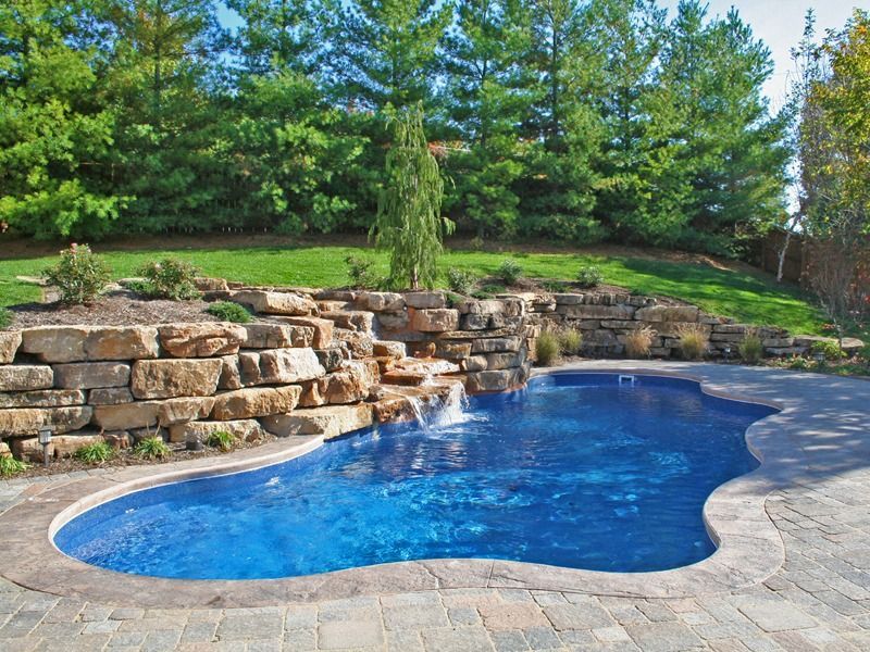 Blue backyard swimming pool beside stone retaining wall and green landscaped trees