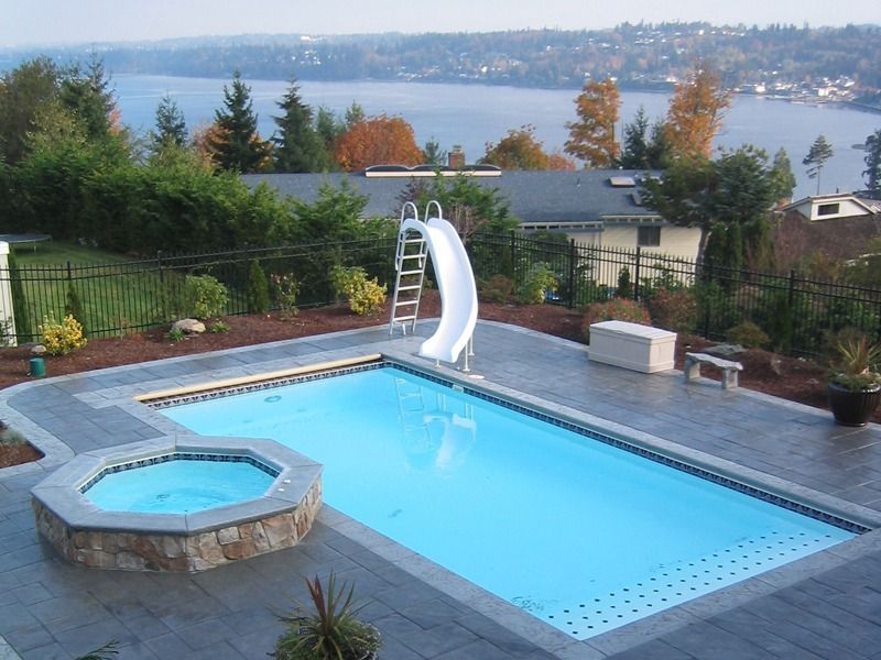 Backyard pool with hot tub, waterslide, and lake view beyond a deck and greenery