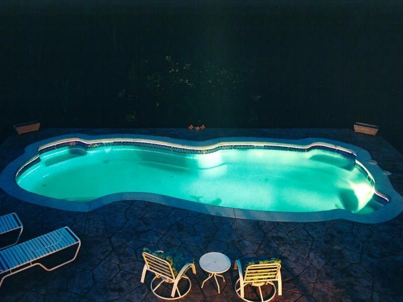 Lit swimming pool glowing turquoise at night, with patio chairs and table nearby.
