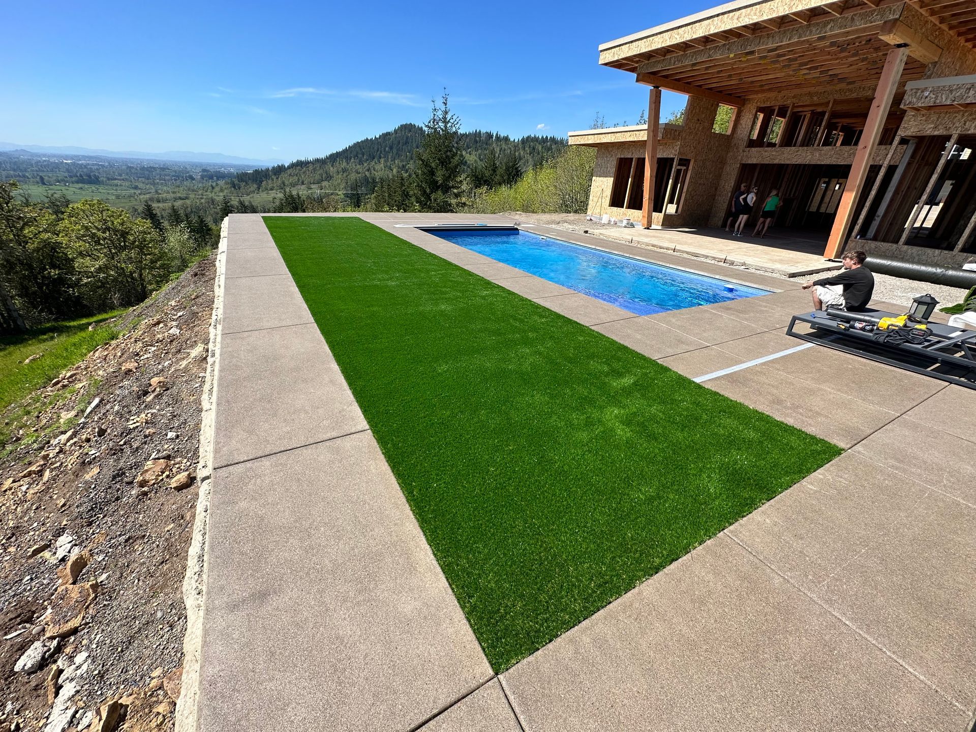Patio with a long green lawn strip beside a blue pool and modern house overlooking a scenic valley