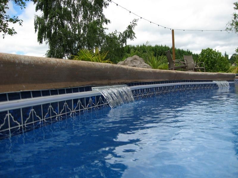 Backyard pool with blue water cascading over a tiled edge under a cloudy sky