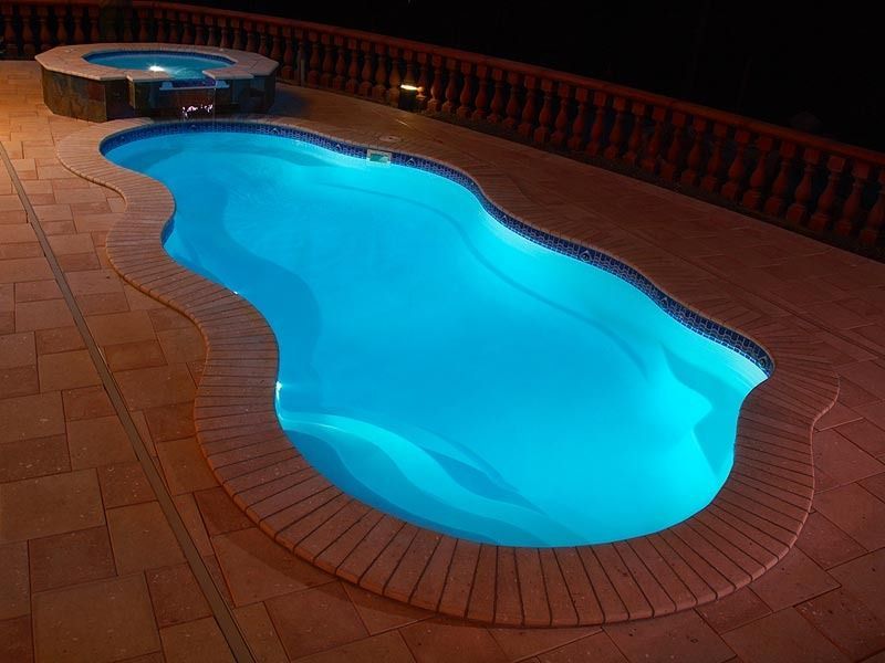 Blue swimming pool with a curved shape, lit at night on a terracotta tile patio.