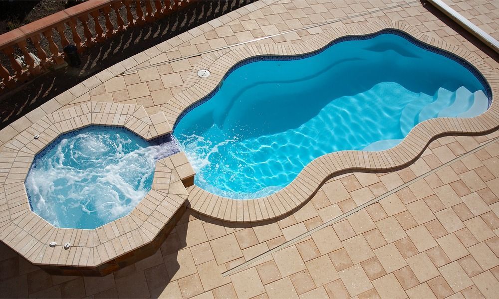 Two connected backyard pools, one small round spa and one larger blue pool on beige tile decking