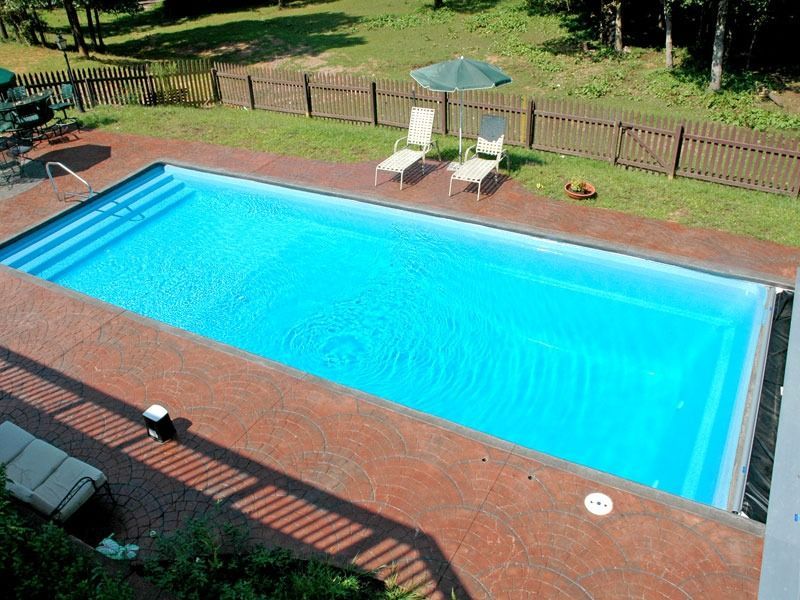 Outdoor swimming pool with bright blue water, brick deck, lounge chairs, and a small umbrella in a fenced yard
