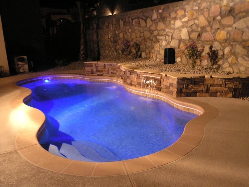 Blue-lit backyard pool beside a stone patio and wall at night
