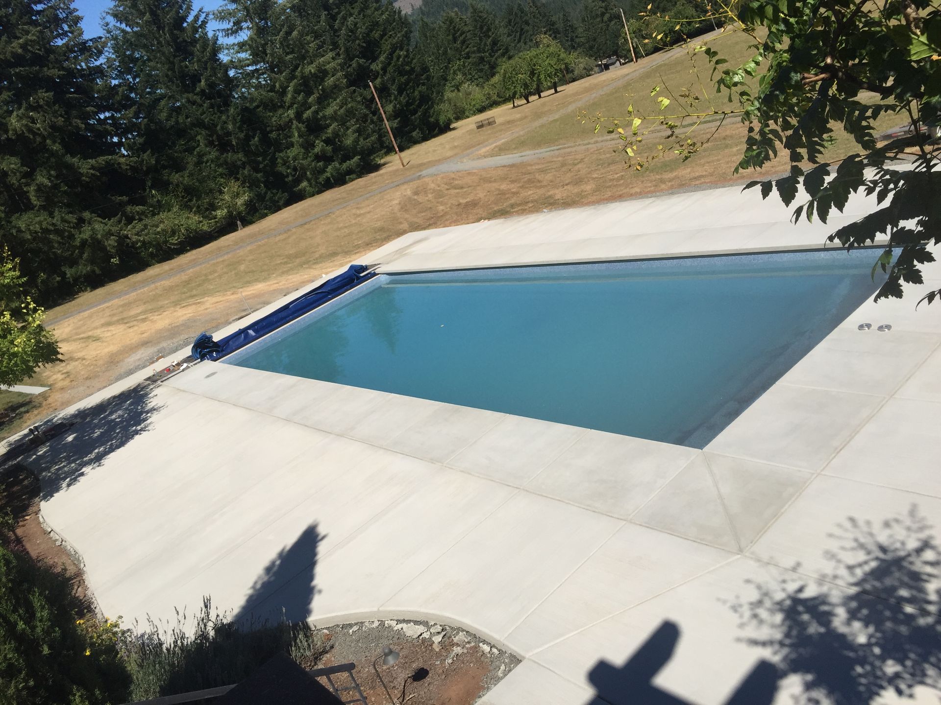 Empty outdoor swimming pool with blue cover, surrounded by concrete patio and trees on a sunny hillside.