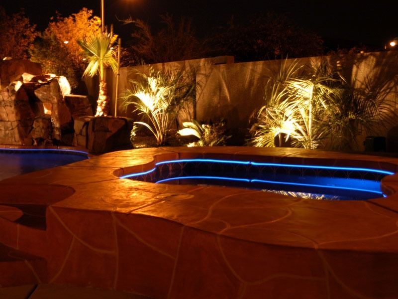 Nighttime pool with blue-lit spa, stone patio, and illuminated desert landscaping