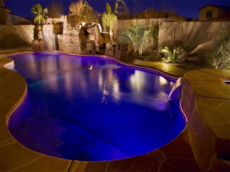 Illuminated backyard swimming pool at night with purple-blue lighting and desert landscaping
