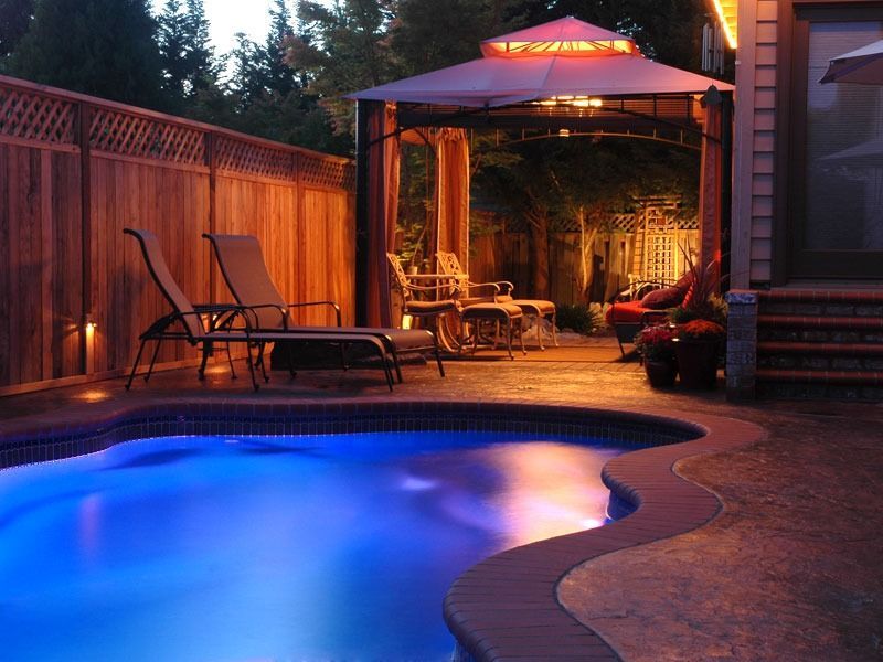 Backyard patio at dusk with glowing blue pool, lounge chairs, and a lit gazebo seating area