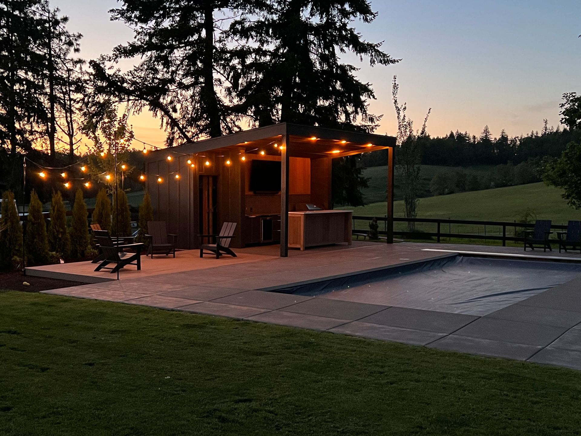 Lit wooden cabin with string lights beside a patio and grassy field at dusk