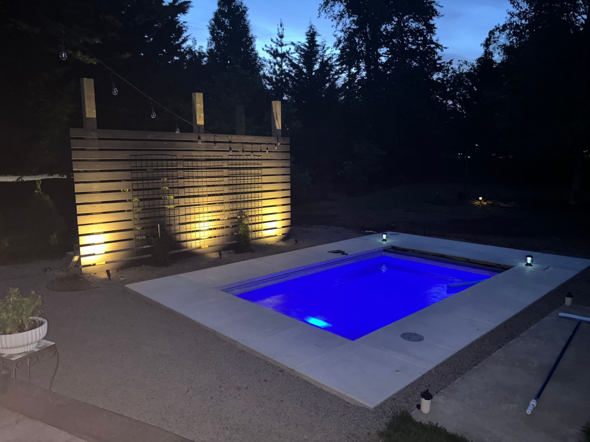 Nighttime backyard with a lit blue hot tub beside a stone wall and trees.