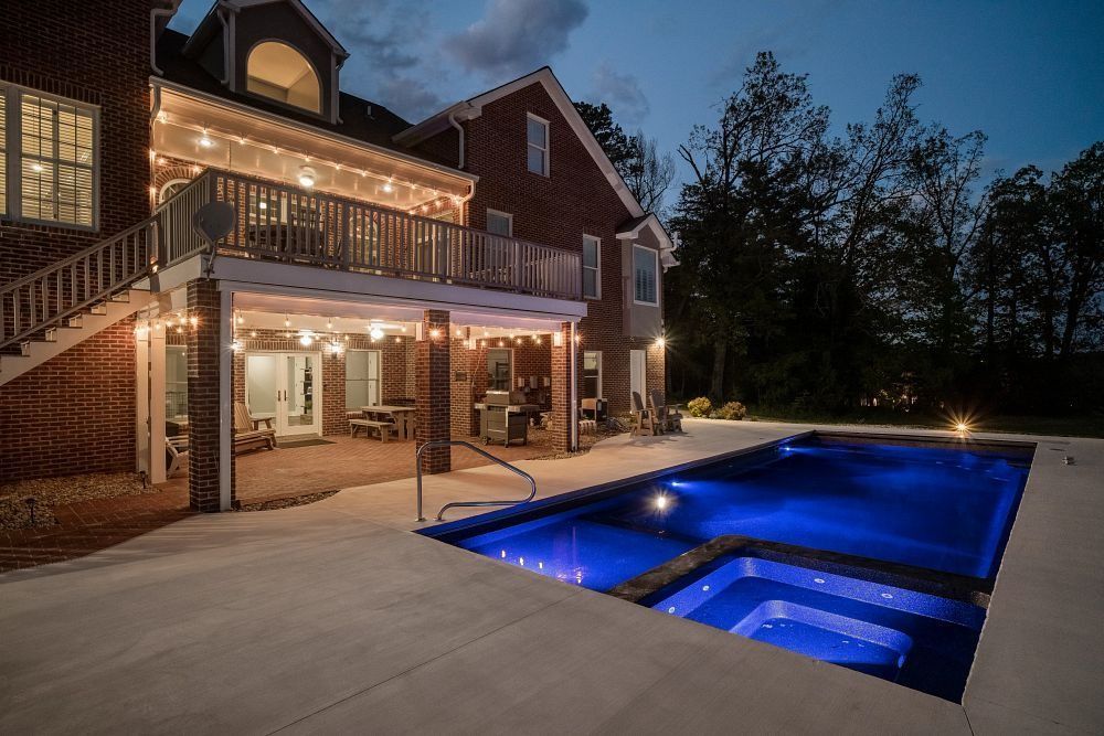 Backyard patio with glowing pool beside a lit two-story house at dusk