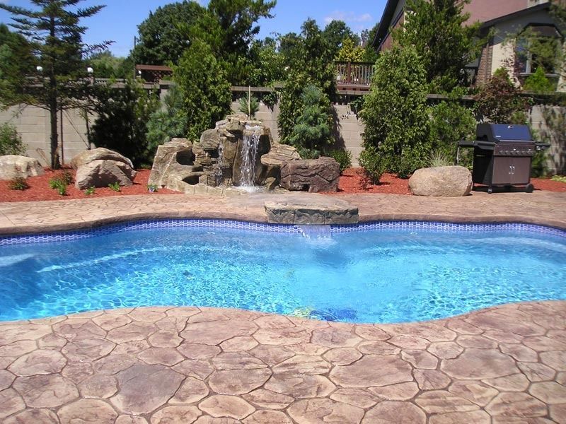 Blue backyard pool with stone deck, waterfall feature, and lush landscaping in sunlight