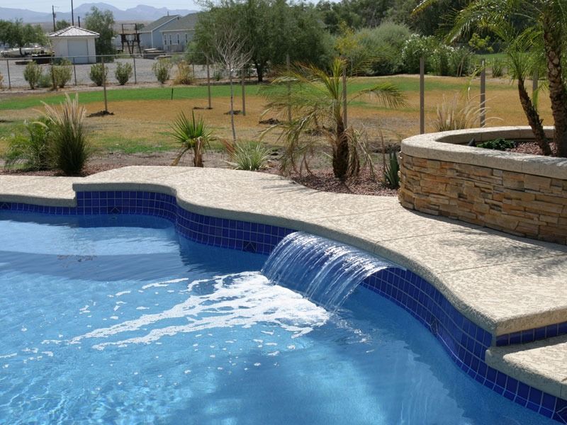 Curved blue swimming pool beside a stone patio and landscaped yard with mountains in the background