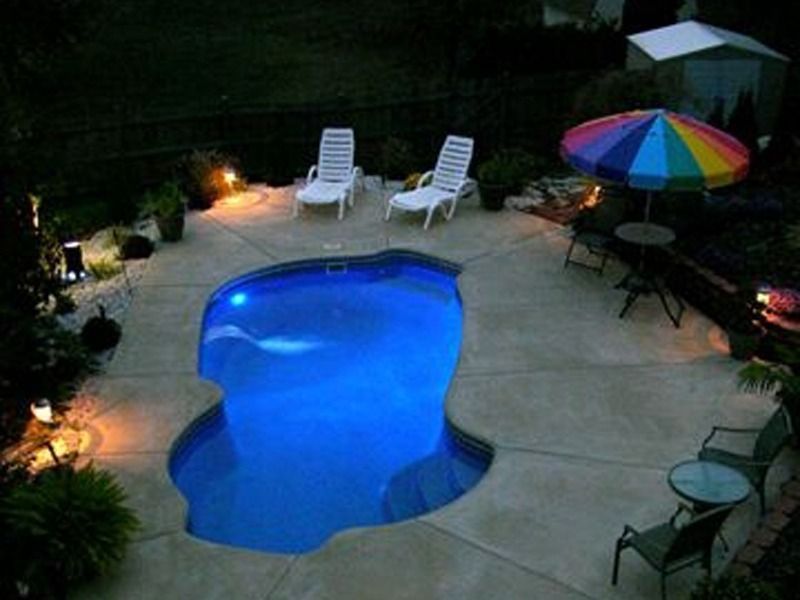 Backyard pool at night with blue water, lounge chairs, patio table, and colorful umbrella