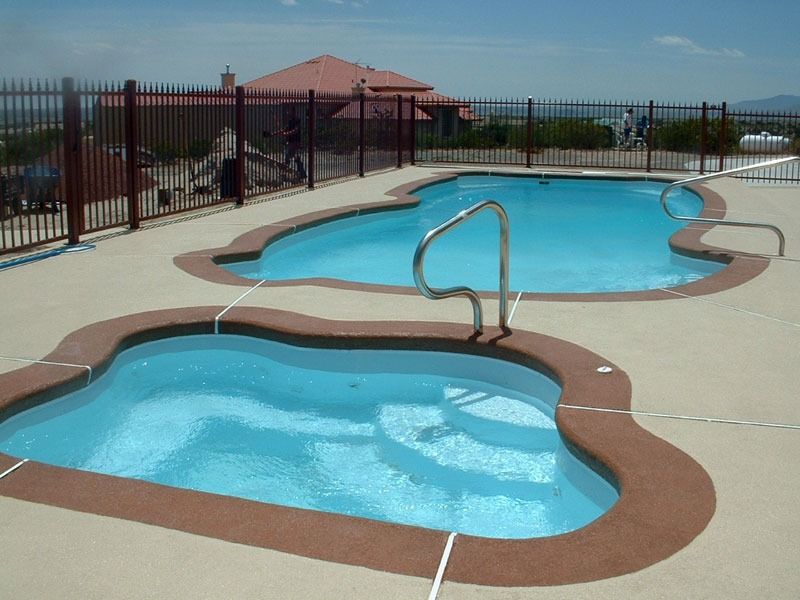 Two small blue swimming pools with a handrail on a sunny rooftop deck, surrounded by a fence.