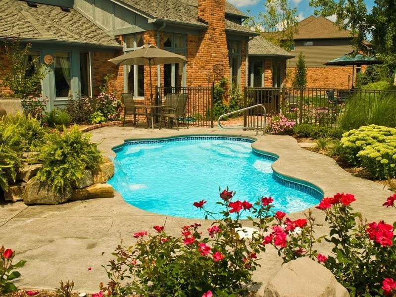 Backyard pool beside a stone house, with patio seating, flowers, and lush greenery.