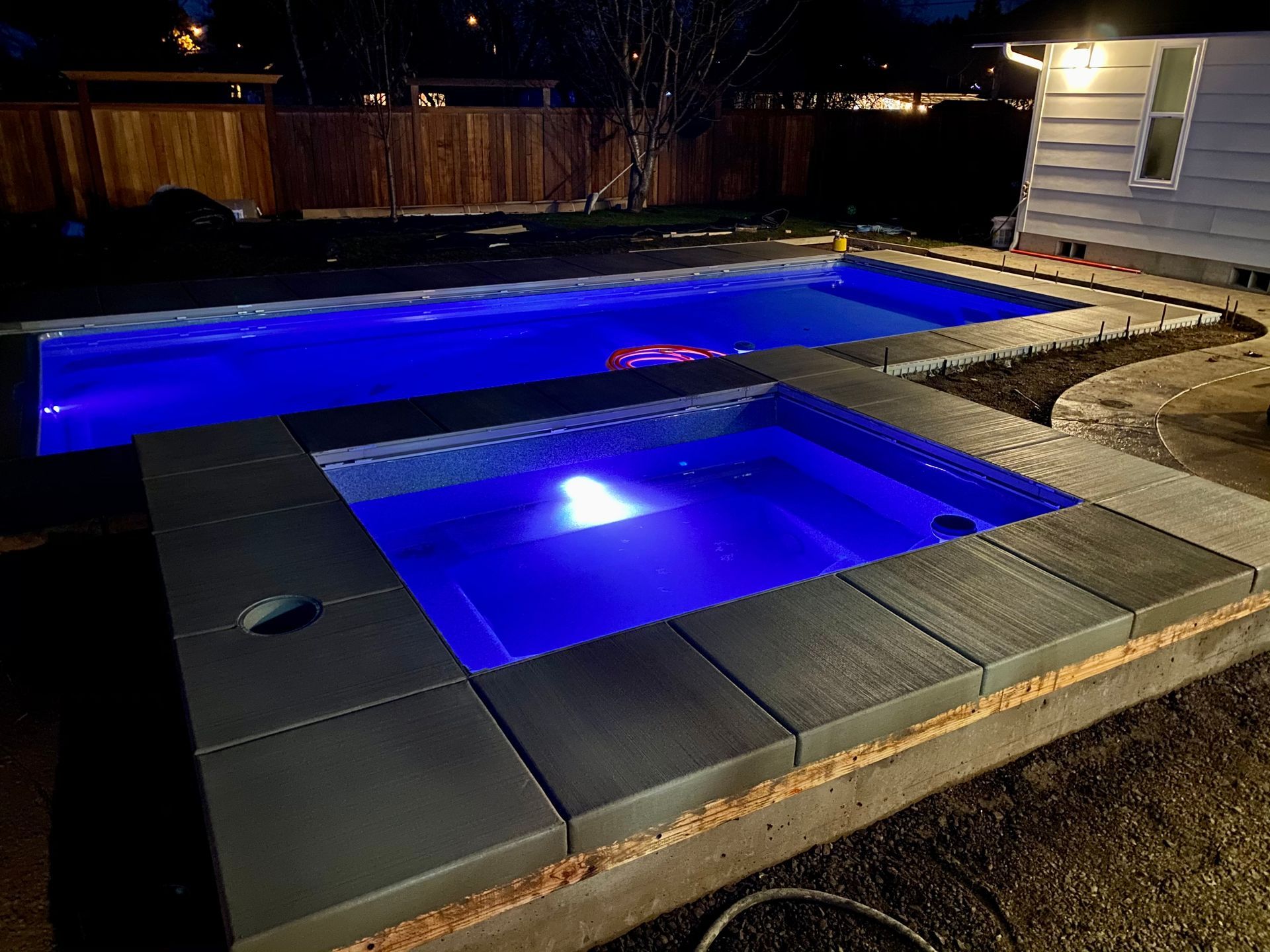 Backyard pool and hot tub glowing blue at night beside a house and fence