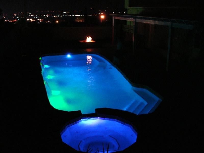 Glowing blue backyard pool at night with a lit fountain and hot tub in the foreground