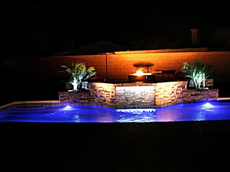 Illuminated backyard pool with blue lighting, stone waterfall feature, and potted palm plants at night