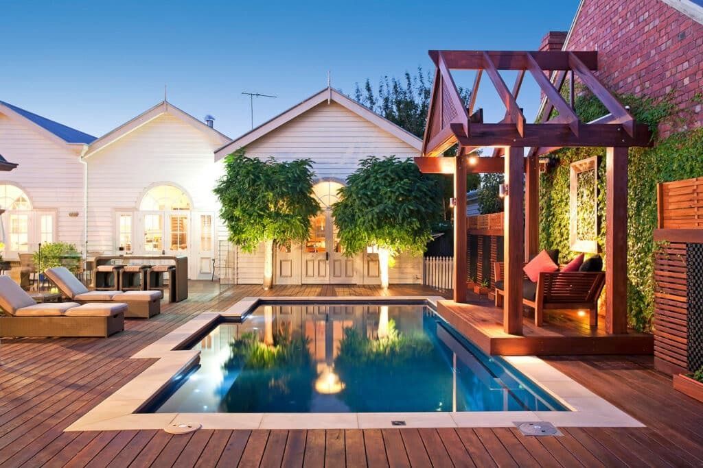 Backyard patio with a swimming pool, lounge chairs, and a lit pergola beside a house at dusk