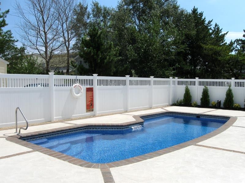 Backyard swimming pool with blue water, white fence, and beige deck under trees