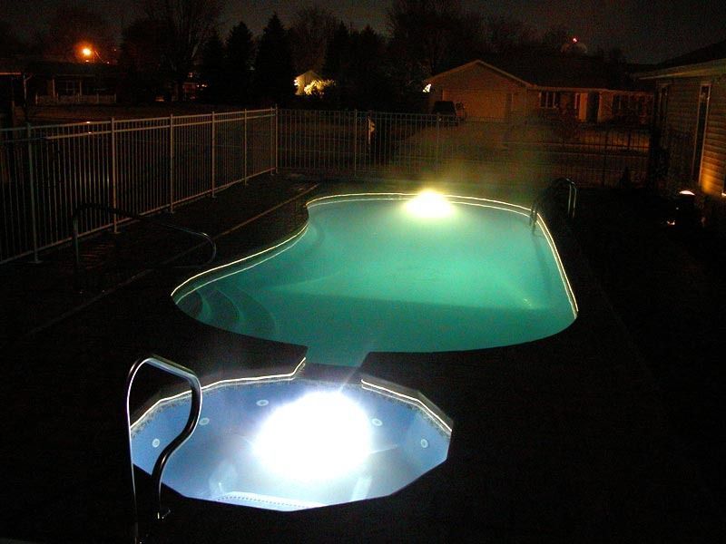 Nighttime backyard pool glowing turquoise and white, with a lit diving board and fence in the background